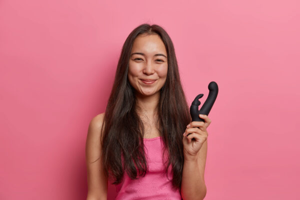 Smilende kvinne i rosa topp holder en svart rabbit dildo foran rosa bakgrunn – illustrasjon av dobbel stimulering og glede ved bruk av rabbitvibrator.