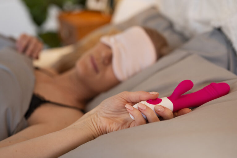 Kvinne hviler i sengen med en liten rabbitvibrator i hånden – illustrerer stillegående nytelse og diskret bruk i rolige omgivelser.