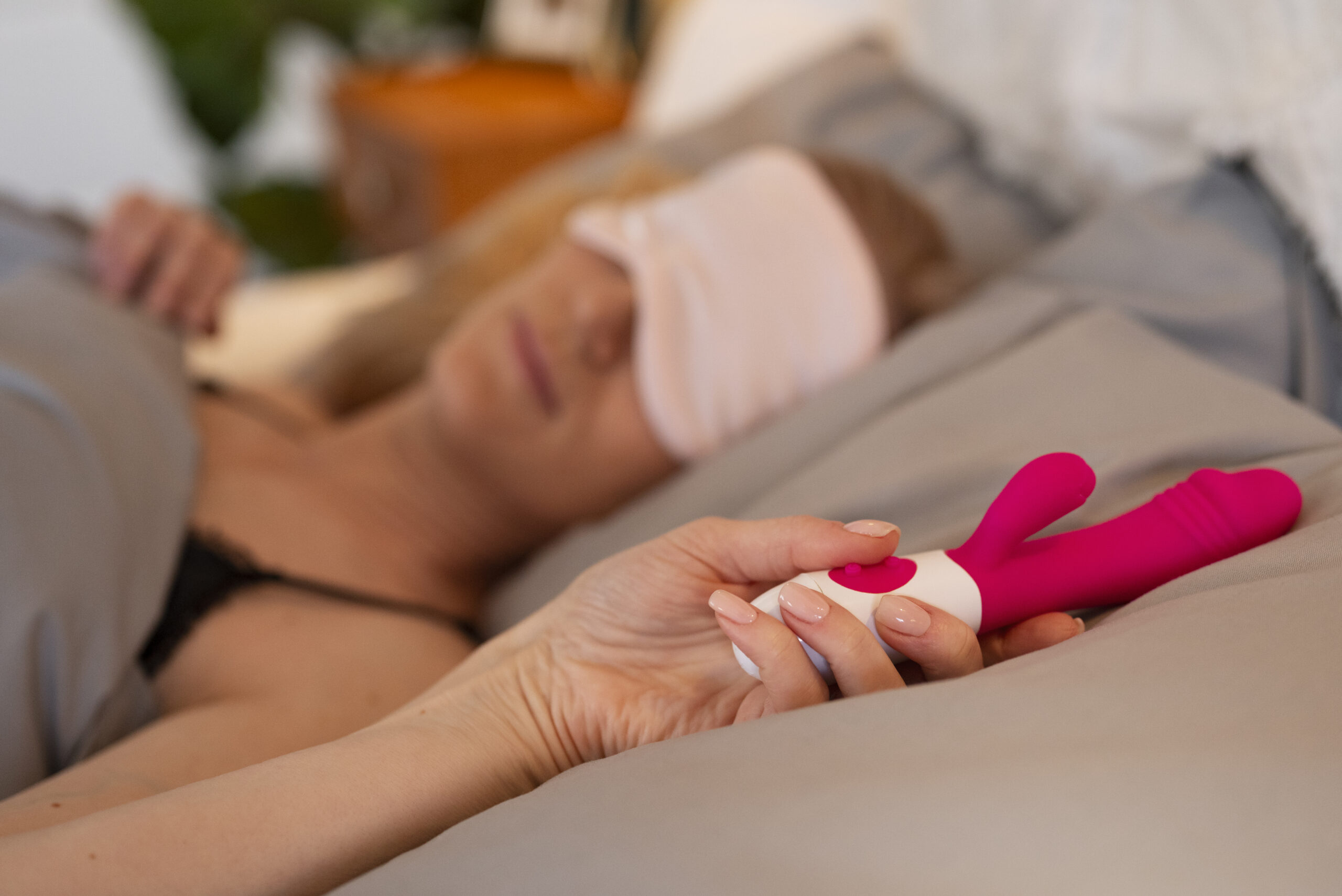 Kvinne hviler i sengen med en liten rabbitvibrator i hånden – illustrerer stillegående nytelse og diskret bruk i rolige omgivelser.