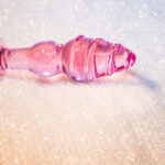 Rosa glassdildo med tydelig base og spiralform, liggende på mykt, hvitt underlag.